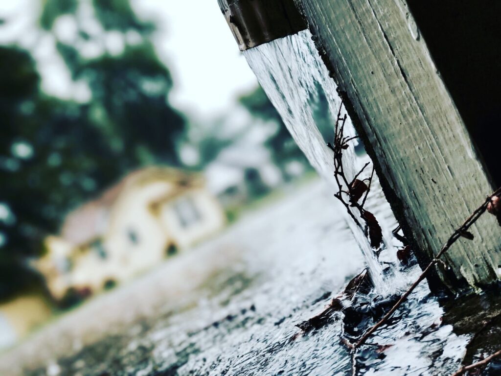 Rainwater pouring hard from a downspout directly onto pavement beside a home's foundation